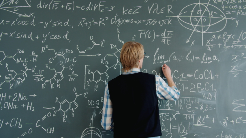 Back view of young man maths teacher writing equations on chalkboard in classroom busy with calculations. Occupation, lifestyle and people concept.