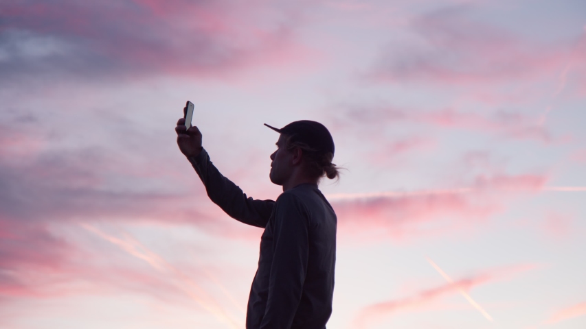 Silhouette of young hipster man make photo or video of pink and purple sunset. Social media influencer or blogger.  Romantic wanderlust concept