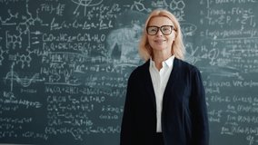 Slow motion of friendly teacher wearing suit and glasses smiling looking at camera standing alone in classroom, blackboard with scientific formulas is in background. - Powered by Shutterstock - Get 15% off with code: PIKWIZARD15