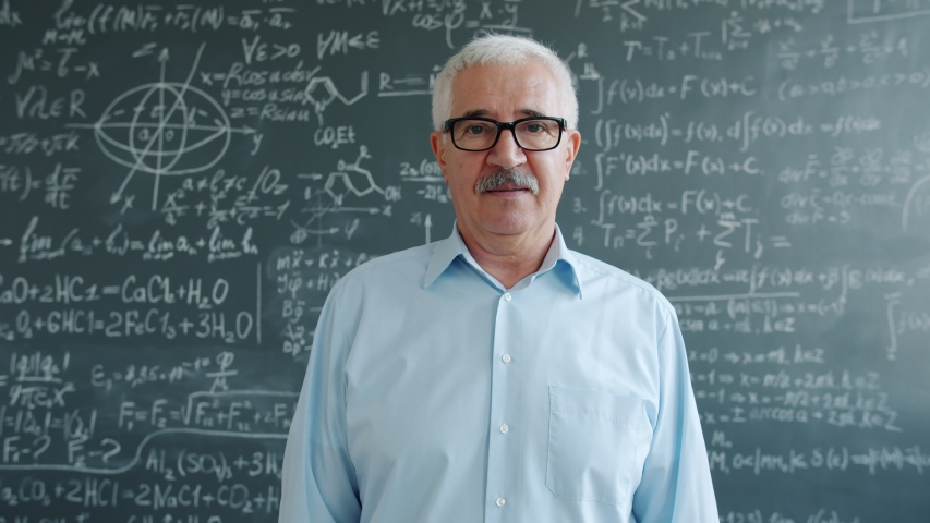 Portrait of smart mature man standing in class with serious face looking at camera, blackboard with formulas is visible in the background. People and science concept.