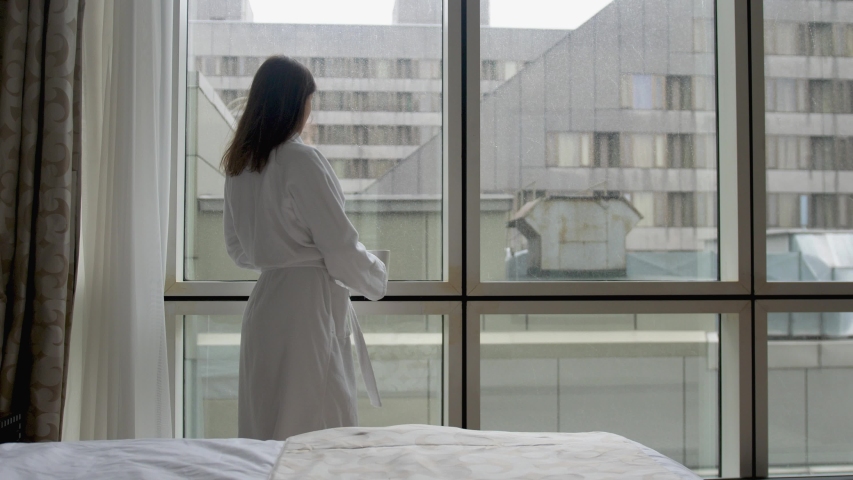 Young woman in white bathrobe stands by the window in hotel apartment with smartphone and cup of coffee