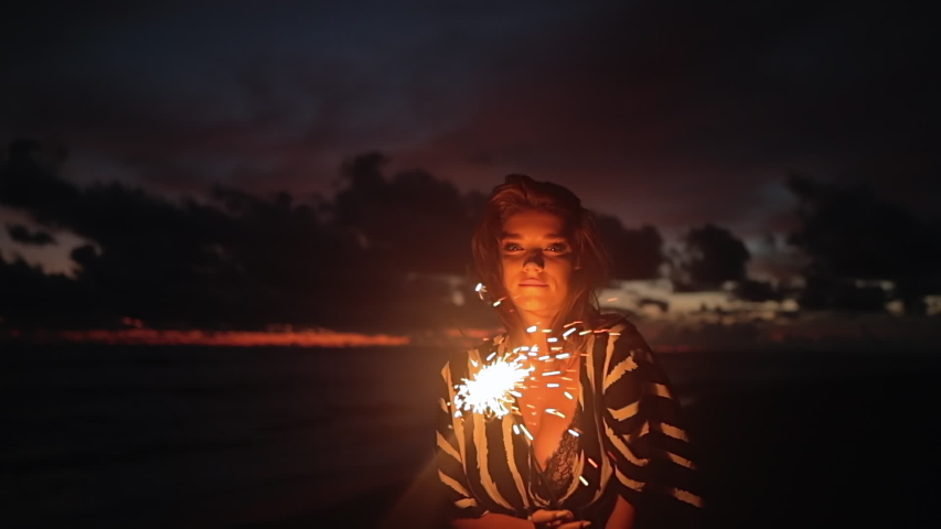 Close up portrait of young beautiful woman at sunset on beach. Pretty girl dancing in with sparkler at sunset in slow motion. Holiday with fireworks. City people travel. Business woman at vacation.