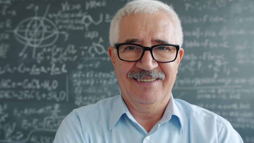 Portrait of happy mature man in glasses looking at camera smiling near chalkboard with formulas written with chalk. Education and positive emotions concept.