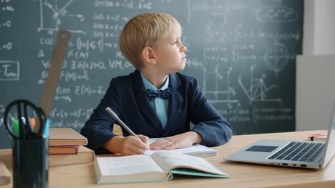 Prodigy kid in suit is taking notes sitting at desk in university classroom with chalkboard covered with formulas in background. Modern education and talanted children concept.