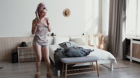 Morning fun background of sofa cushions funny girl dressed in nightwear home slippers, wearing wireless headphones, dancing in living room smiling looking at mobile phone, hair wrapped in bath towel