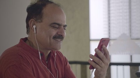 A happy old man using smartphone and earphones to chat with a wife over a video call in a long distance relationship. Smiling Grandpa talking with his son friend working abroad in indoor home setup. 