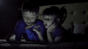 Two young children using a creative painting app on a tablet or mobile phone while lying on the bed. A boy and a girl are busy playing video games on smartphone in the dark. 