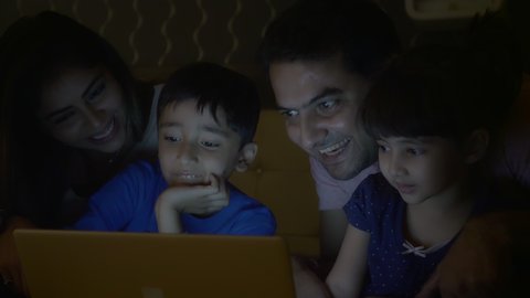 A family of four members including parents and two young siblings are enjoying while using tablet or Laptop in the night. A happy family smiling while watching a movie or playing online video game. 