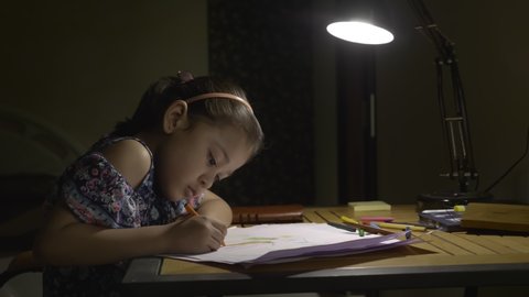 A young girl child is busy drawing on a white paper under a table or desk lamp in the night. A adorable kid studying in a study room during the exams. 