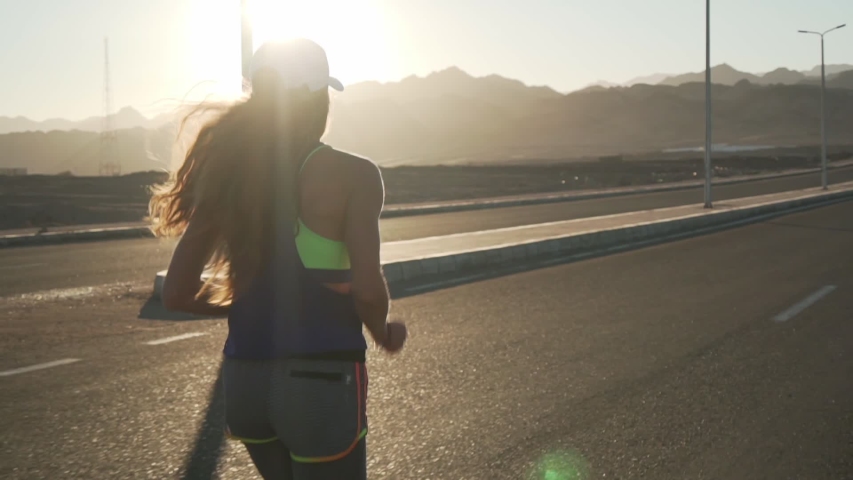 Back view slim girl jogging at highway in mountains at sunset rapid slow motion