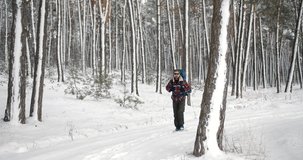 Mature male tourist in warm clothing walking through frosty winter forest with backpack. Bearded man with sport equipment hiking during snowy days.