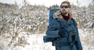 Mature bearded man in winter coat, gloves and eyewear standing among snow-covered pine trees. Serious hiker with big backpack and binoculars enjoying winter season