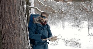 Mature bearded man wearing in hiking equipment looking at map to find easy way through snowy coniferous forest. Male tourist in sporty eyeglasses standing near trees to orientate around