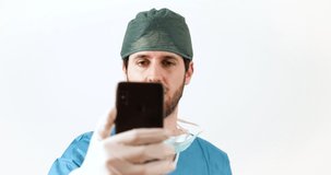 Doctor, in blue uniform, is taking a selfie video with his smart phone. White background.