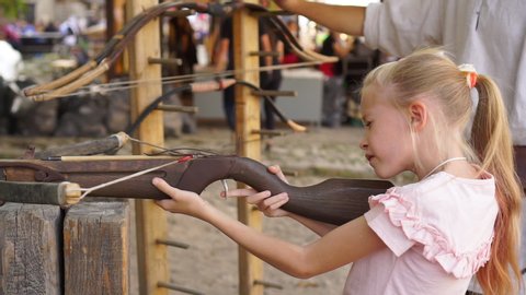 Blond child girl aims and shoots with crossbow at open air amusement park medium shot handheld
