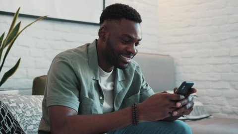 Close up of amazed young african-american man use phone smiles sit on sofa in home online relax work cell phone app authentic bills sunset guy close up mobile smartphone slow motion portrait handsome