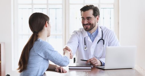 Smiling male professional doctor plastic surgeon handshakes female patient agree on surgery. Happy physician in white uniform shake hand of woman at medic checkup visit, medical service trust concept
