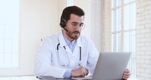 Professional male doctor in white medical coat and headset making conference call on laptop computer, consulting distance patient online in video chat, explain treatment by webcam concept