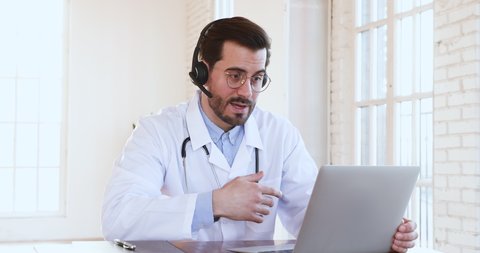 Professional male doctor in white medical coat and headset making conference call on laptop computer, consulting distance patient online in video chat, explain treatment by webcam concept