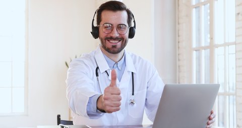 Friendly smiling professional male medic doctor in headset makes conference video call on laptop  consult patient look at camera, shows thumbs up recommend telemedicine online chat service app concept