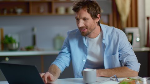Smiling businessman getting good news on laptop computer at home workplace. Business man sitting at table at home office. Happy man using on laptop at kitchen in slow motion.