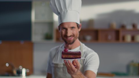 Smiling male baker putting cherry on cake at workplace. Young confectioner finishing dessert on plate at kitchen. Portrait of chef man decorating cake and smiling at camera on kitchen in slow motion