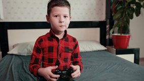 Cute boy in black red shirt is playing video game, holding joystick, having an emotional time. Close up. Throws console on bed