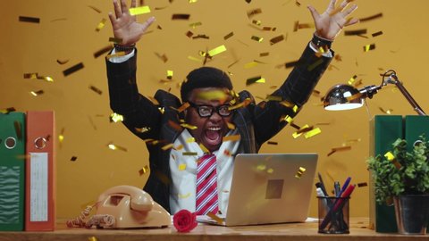 Shocked happy black businessman winning online bets company finance enjoying victory celebrating under confetti rain working in his office.