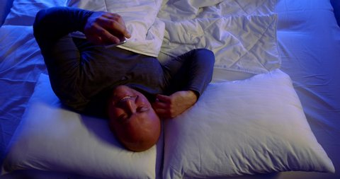 a bald man in dark pajamas is lying on white bed linen. he holds a smartphone, laughs a lot, covers his eyes with hand. the view from the top. blue bottom light
