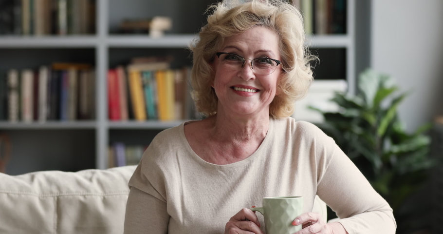 Happy beautiful older middle aged woman look at camera hold tea cup drink coffee sit on sofa alone at home, senior adult retired lady grandma. Dental smile pose for portrait on couch in living room Royalty-Free Stock Footage #1047036799