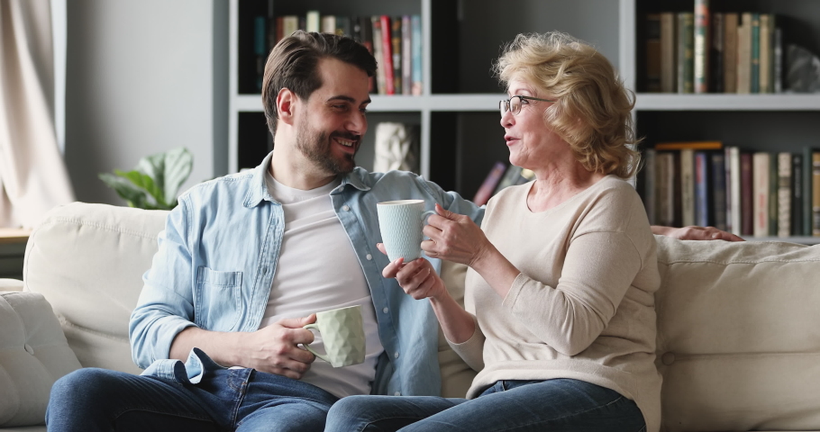 Happy 2 two generations family drinking tea having conversation sit on sofa at home. Older mature mother talking to young adult grown son share news on couch in living room together Royalty-Free Stock Footage #1047036823