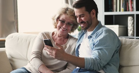 Smiling young adult man grownup son showing funny social media photos, enjoying using smart phone mobile apps sit on sofa with older mature parent. Mom having fun laughing looking at cellphone screen