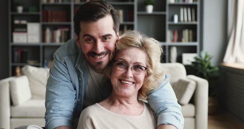Smiling 30s young adult man grown handsome son looking at camera hugging old mature 60s mom expressing love and care embracing cuddling on mothers day concept. 2 generations family closeup portrait