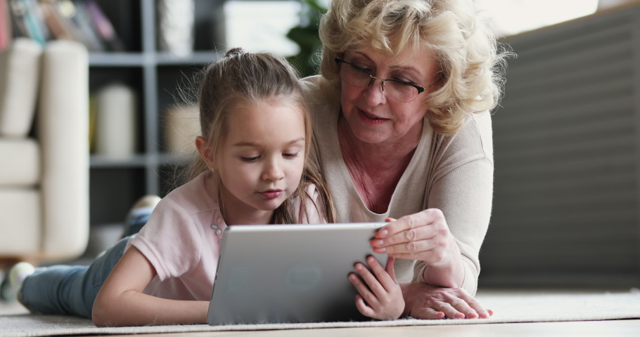 Old mature grandma nanny teaching cute small preschool granddaughter learning reading ebook on digital tablet. Two generation family grandparent and grandchild using pad device tech on floor at home Royalty-Free Stock Footage #1047036844