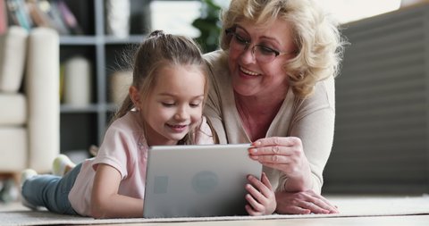 Old mature grandma nanny teaching cute small preschool granddaughter learning reading ebook on digital tablet. Two generation family grandparent and grandchild using pad device tech on floor at home
