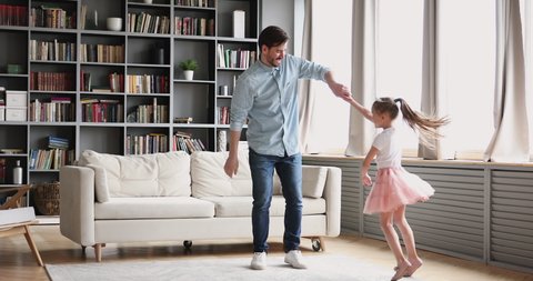Affectionate loving young dad teaching dancing with cute small child. Daughter princess wear crown skirt having fun in modern living room playing ball together enjoy sweet funny activity at home.