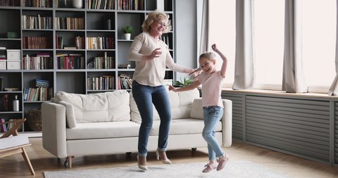 Funny small girl granddaughter dance with old grandma babysitter in living room. Happy two 2 generations active family senior nanny and cute energetic grandkid  enjoy time together at home
