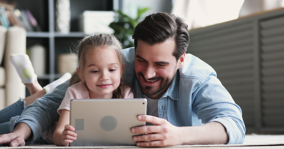Happy family adult parent dad and small daughter having fun using digital tablet lying on floor at home. Cute child girl learning technology talking with father teaching kid look at pad screen at home | Shutterstock HD Video #1047036883