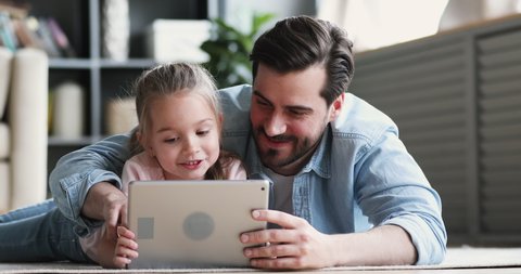 Happy family adult parent dad and small daughter having fun using digital tablet lying on floor at home. Cute child girl learning technology talking with father teaching kid look at pad screen at home