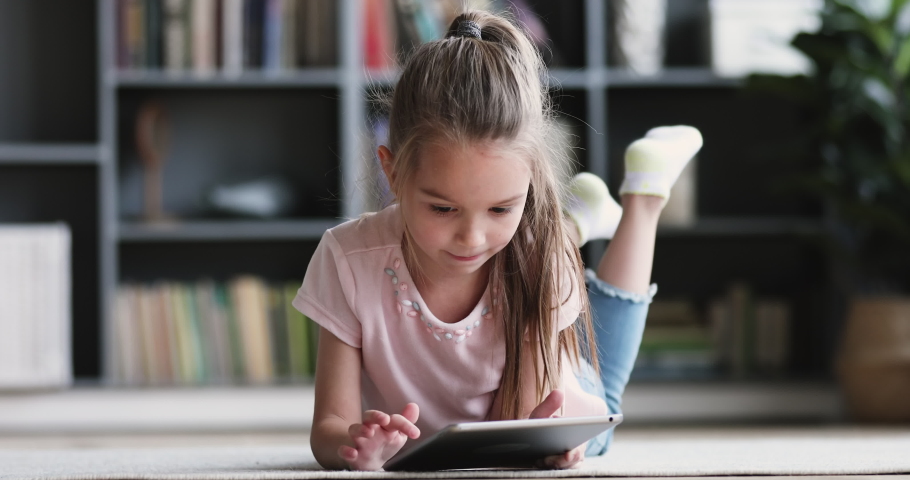 Curious cute preschool kid girl using digital tablet technology device lying on carpet floor alone. Small child hold pad computer surfing internet play game at home. Children tech addiction concept | Shutterstock HD Video #1047036895