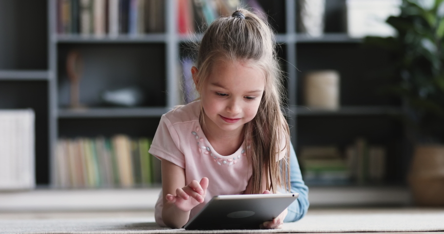 Curious cute preschool kid girl using digital tablet technology device lying on carpet floor alone. Small child hold pad computer surfing internet play game at home. Children tech addiction concept | Shutterstock HD Video #1047036895