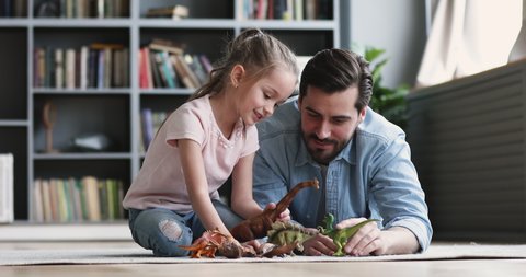 Smiling adult daddy and cute small kid daughter having fun playing dinosaurs toys spend leisure time together at home. Happy single father enjoy funny game with little preschooler child girl on floor