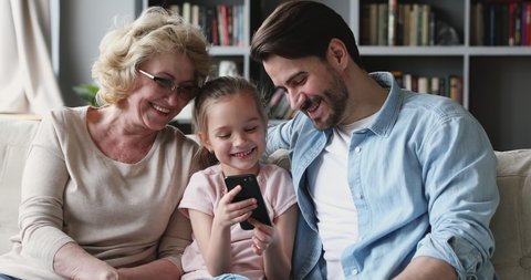 Happy multigenerational 3 age generation family using smart phone mobile gadget sit on sofa, cute small kid granddaughter recording funny video, having fun with young adult dad and old grandma at home