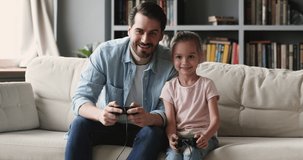 Happy father plays video games with cute kid daughter, family gamers adult parent dad and child girl hold. Enjoy activity having fun winning videogame together sit on sofa at home