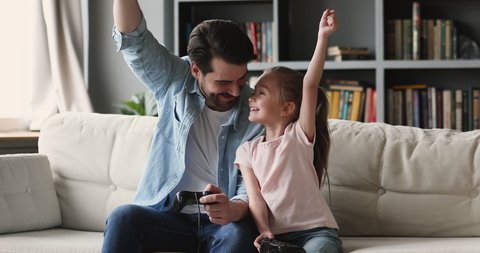 Happy father plays video games with cute kid daughter, family gamers adult parent dad and child girl hold. Enjoy activity having fun winning videogame together sit on sofa at home
