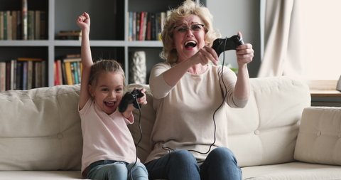 Funny excited two generations family gamers play video game at home. Happy old grandmother and cute child girl granddaughter win videogame hug celebrate victory sit on sofa