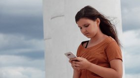 Beautiful brunette teen girl in a brown ochre t-shirt looking at the phone against the sky and the colonnade