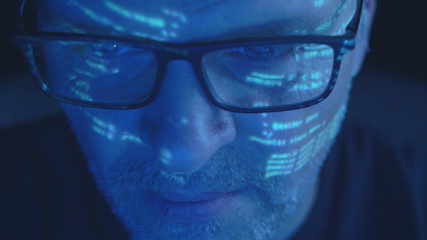 Closeup on the eye movement of a man looking into the screen with data reflecting on his face at night.