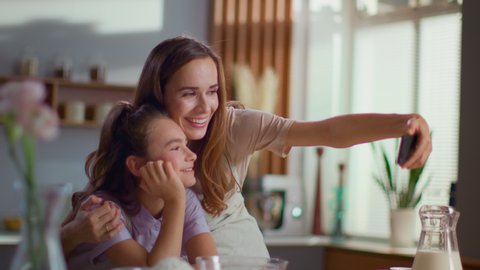Happy mother and daughter taking selfie photo on smartphone at domestic kitchen. Smiling girl and woman using cellphone for self portrait together at home in slow motion