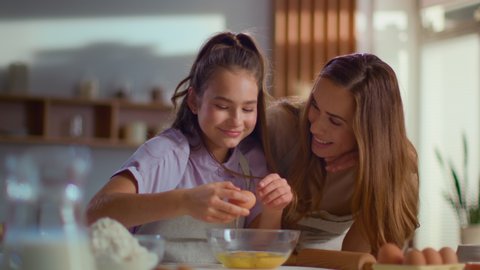 Smiling mother and daughter cracking egg into mixing bowl on kitchen in slow motion. Family preparing dough for cookies together on domestic kitchen. Mom and child cooking together at home